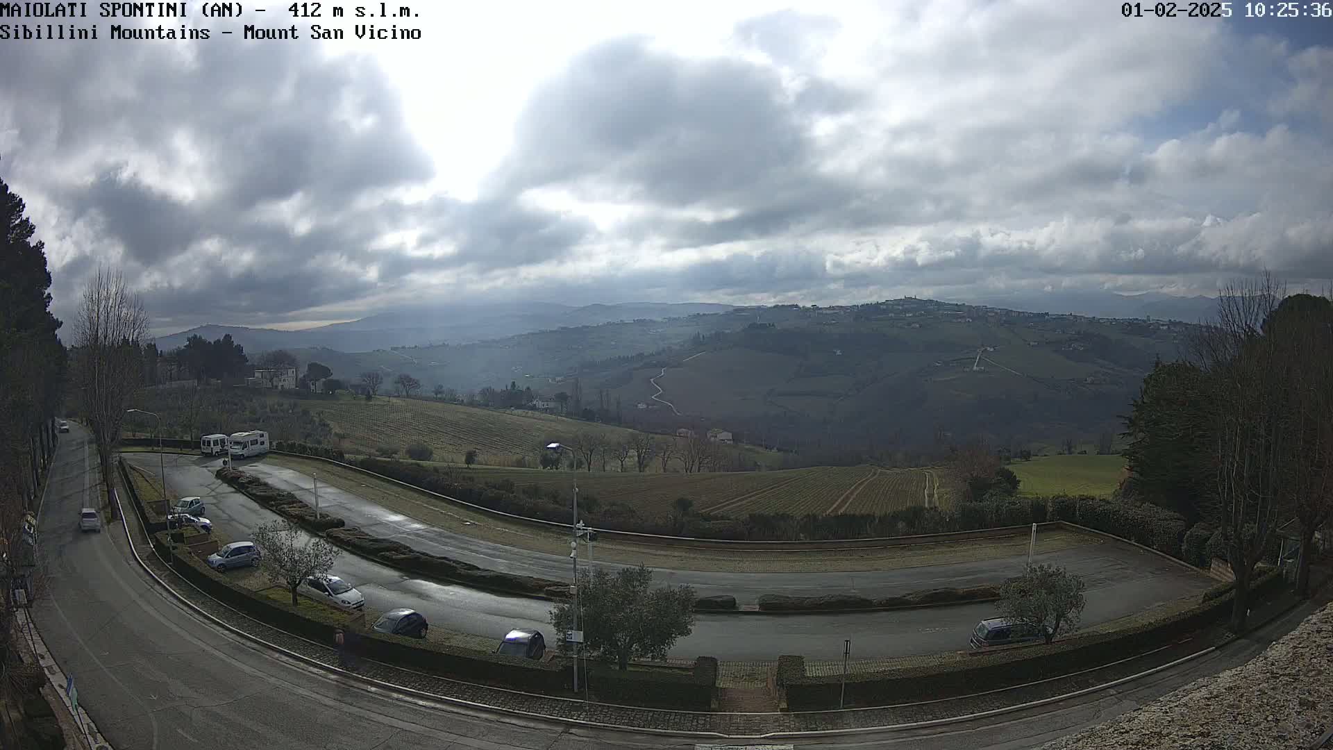 A winding road curves through a parking lot overlooking a hilly landscape under a cloudy sky.