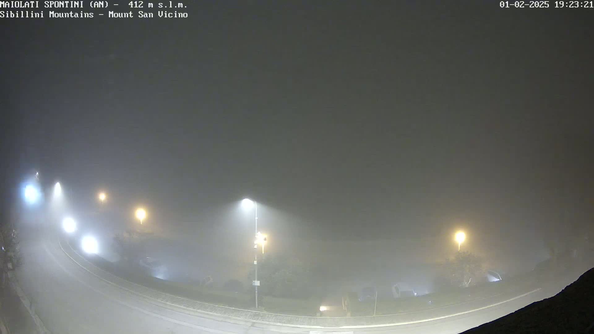 A foggy night scene shows a curving road with illuminated streetlights, and some dimly lit buildings are barely visible in the thick fog.