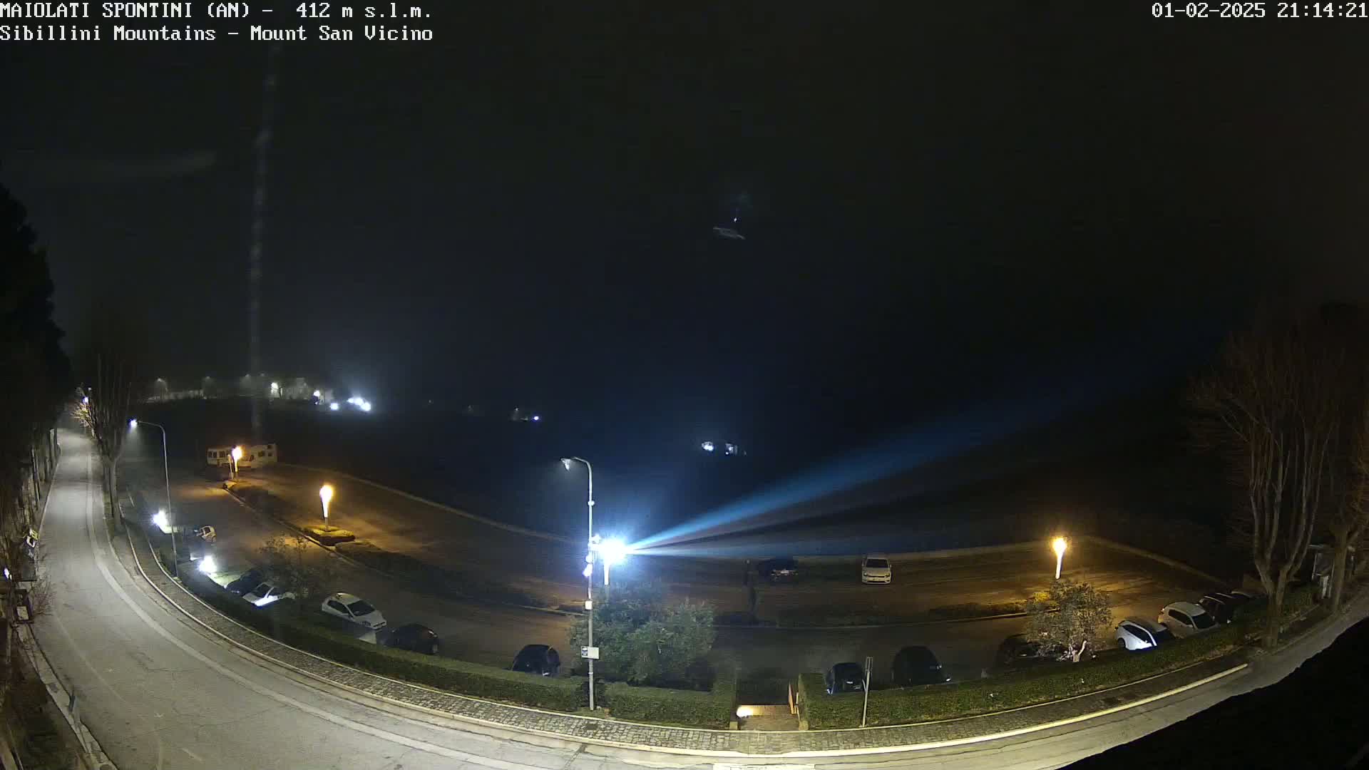 A nighttime, foggy view of a curved road with parked cars and streetlights, illuminated by a bright beam of light projected across a mostly empty parking lot.