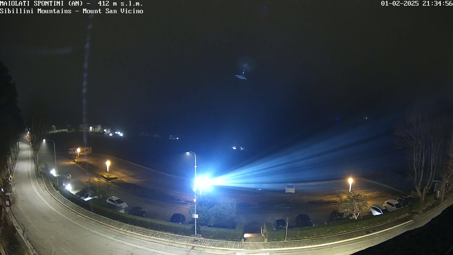 A nighttime, foggy view of a parking lot and curved road illuminated by streetlights and a powerful spotlight projecting beams of light.