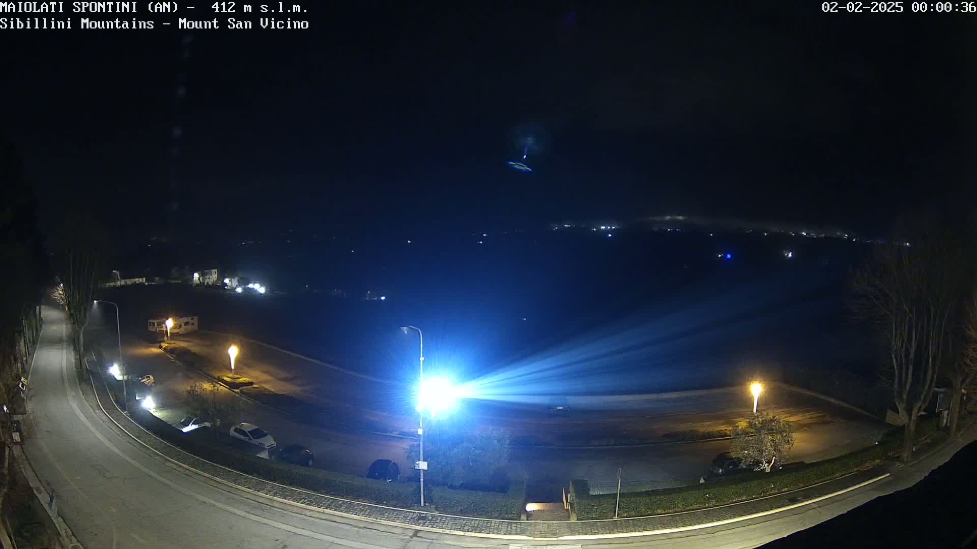 A nighttime, high-angle view shows a road curving through a parking lot with several cars, illuminated by bright streetlights, under a mostly clear, dark sky.