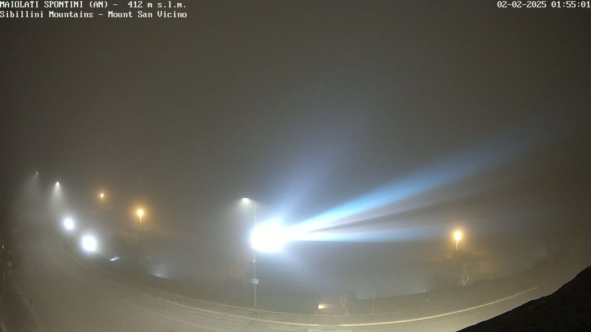 A foggy night scene shows a curved road with several streetlights illuminating the fog, creating bright beams of light.