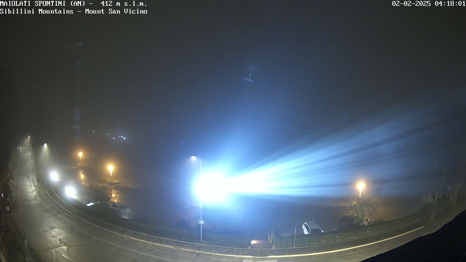 A bright light cuts through a foggy night, illuminating a curved road and parked cars in a dimly lit area.