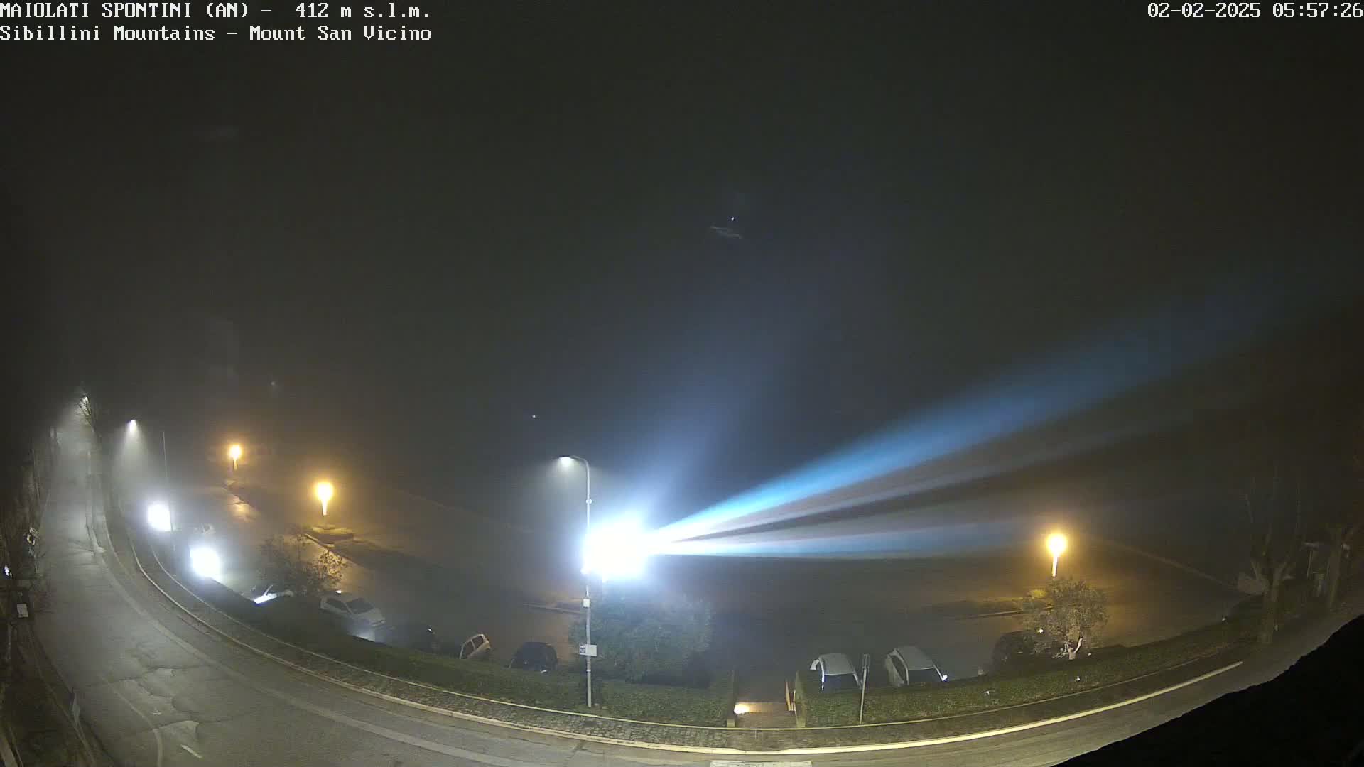 A foggy night scene shows a curved road with parked cars and illuminated by streetlights, with a bright beam of light cutting across the fog.