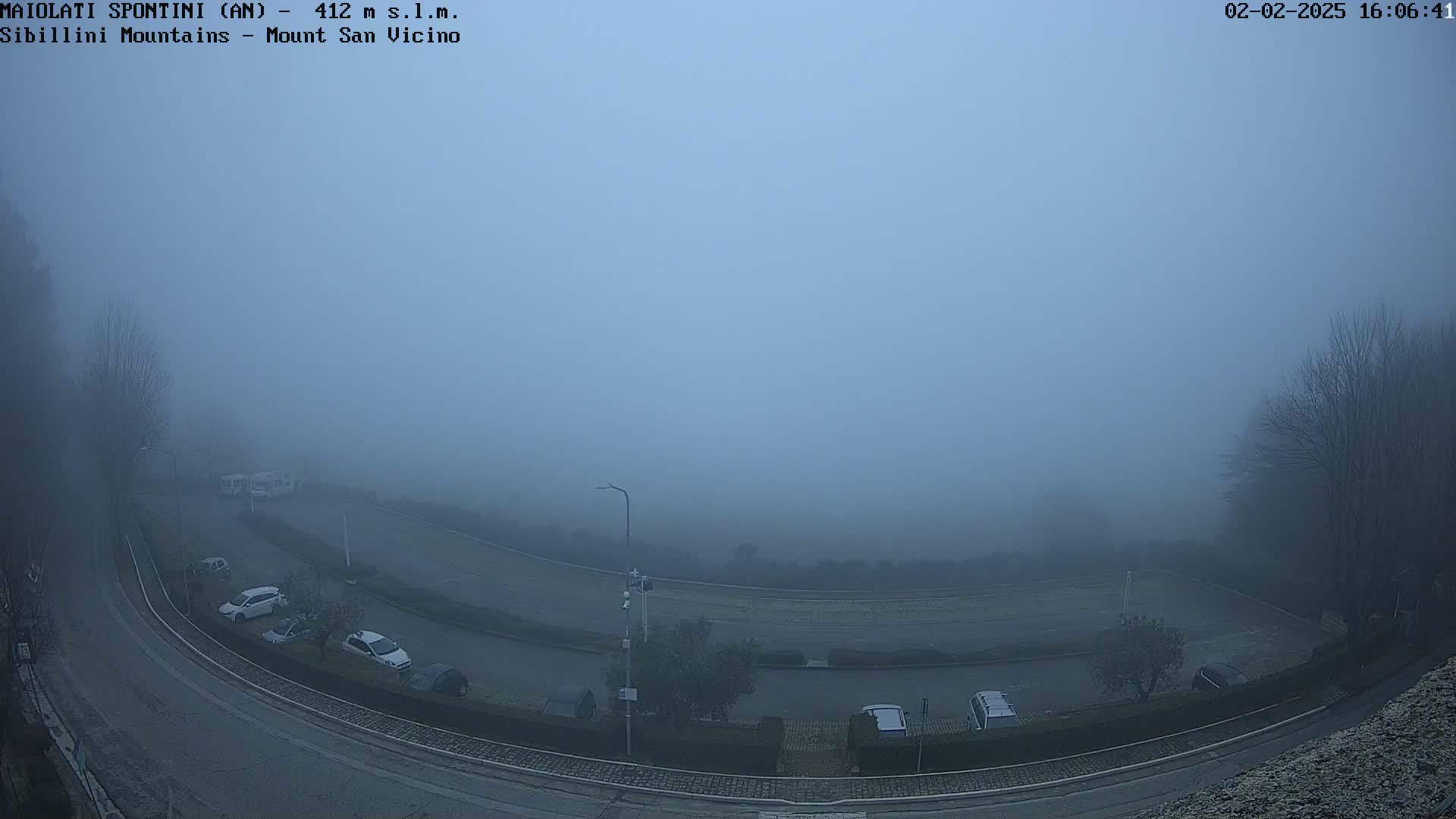 A foggy, high-angle view shows a curved road with parked cars and a few vehicles driving, surrounded by low visibility.