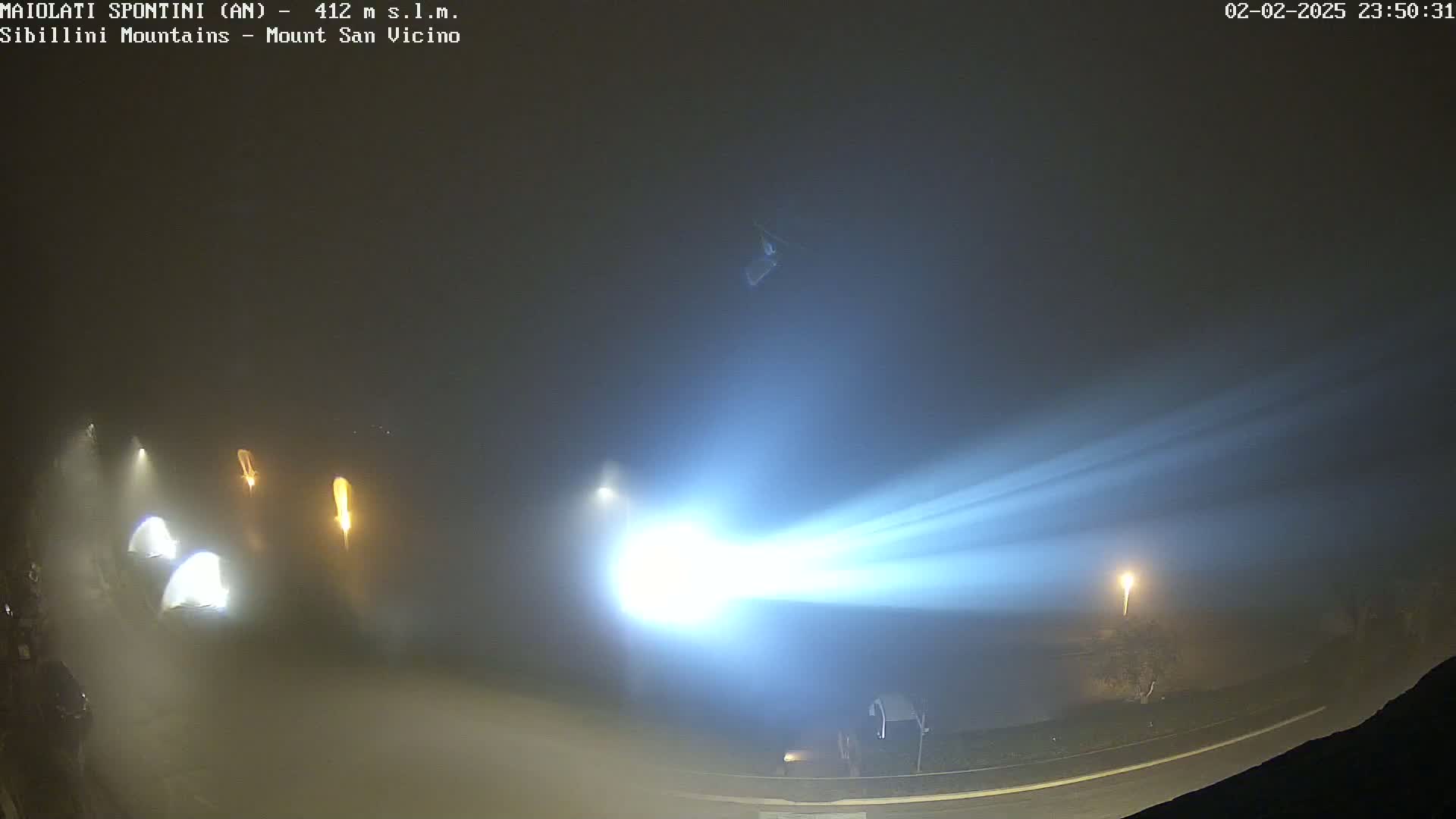 A bright beam of light cuts through thick fog at night, illuminating a road with several vehicles and streetlights visible in the distance.