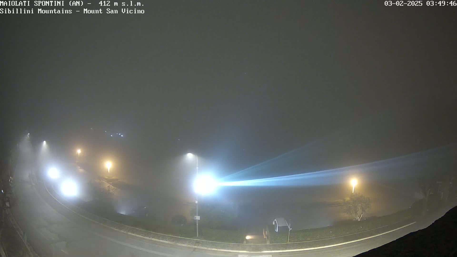 A foggy night scene shows a curved road with illuminated streetlights and some vehicles, all under a dense fog.