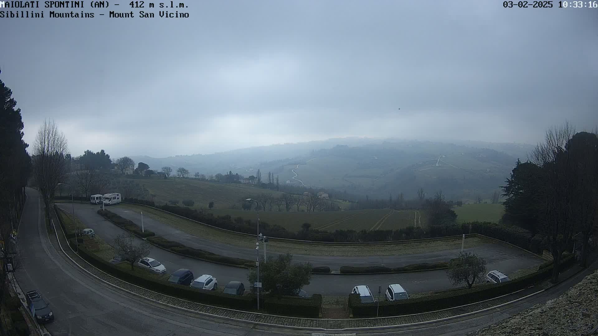 A parking lot with several cars is situated on a hillside overlooking a hazy, overcast valley with rolling hills and farmland.