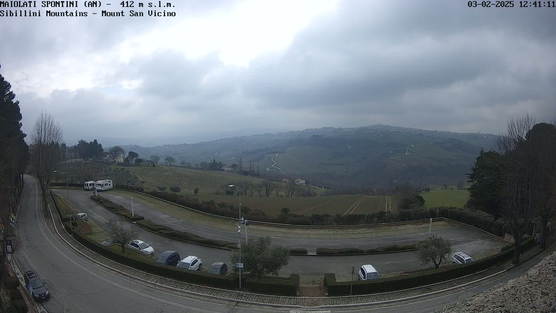 A parking lot with several cars and camper vans overlooks a hilly landscape under an overcast sky.