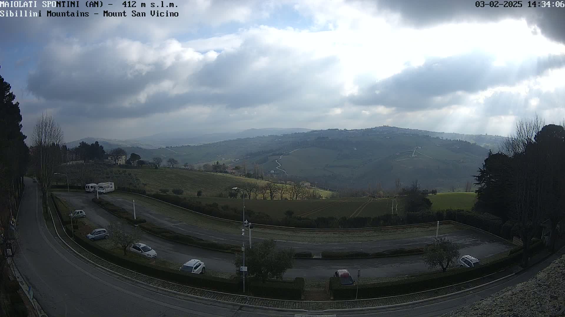 A mostly cloudy day reveals a view of rolling green hills and a parking lot with several cars, all overlooked by a winding road.