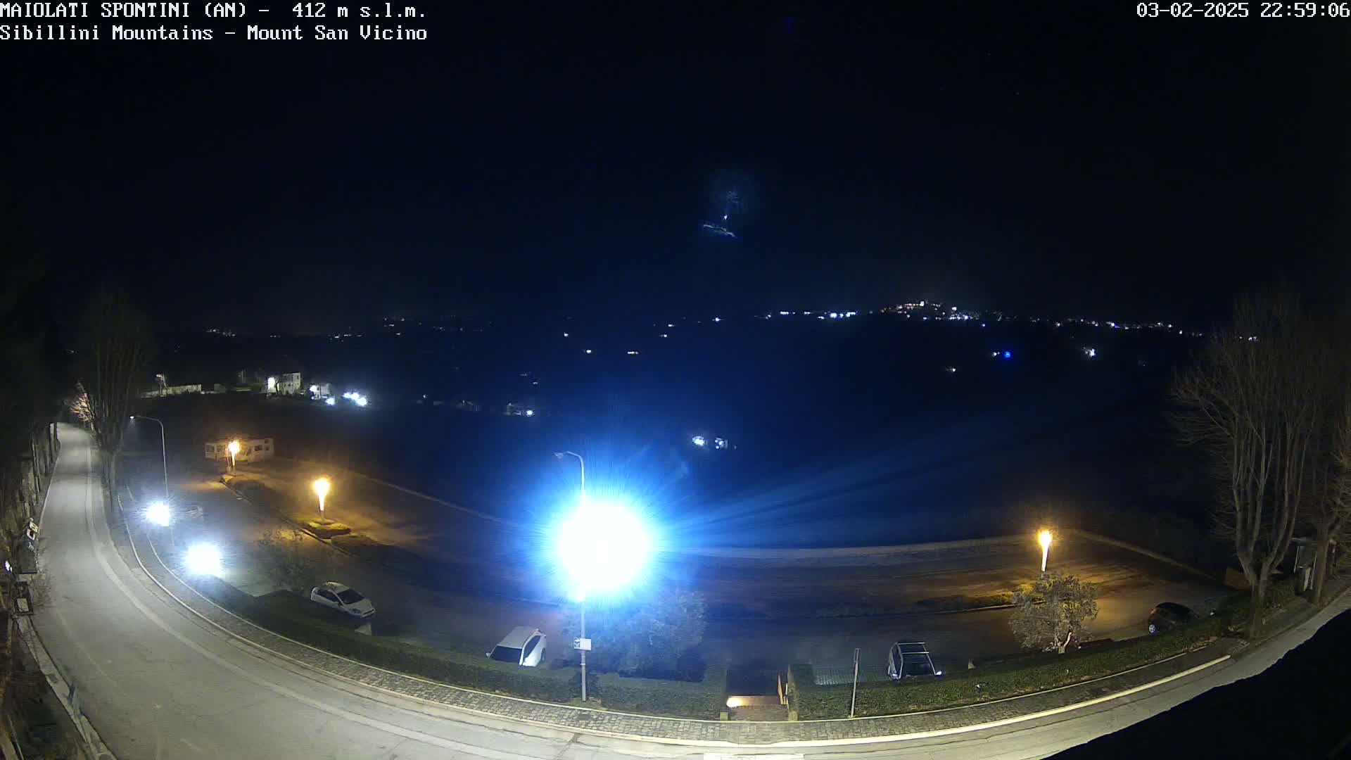 A nighttime, overhead view of a curving road illuminated by streetlights, with parked cars and a distant, dimly lit town under a clear, dark sky.