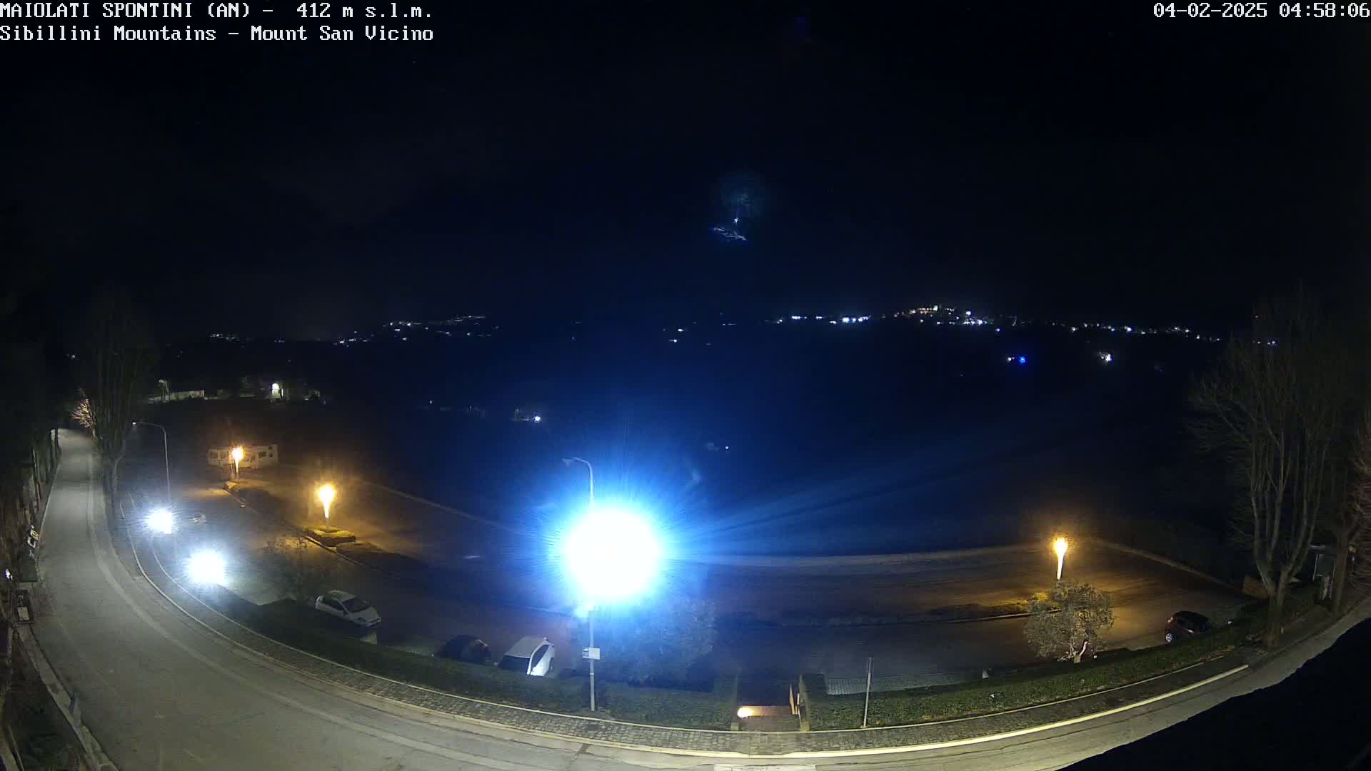 A nighttime, overhead view of a curved road with parked cars and illuminated streetlights, overlooking a distant town under a clear, dark sky.