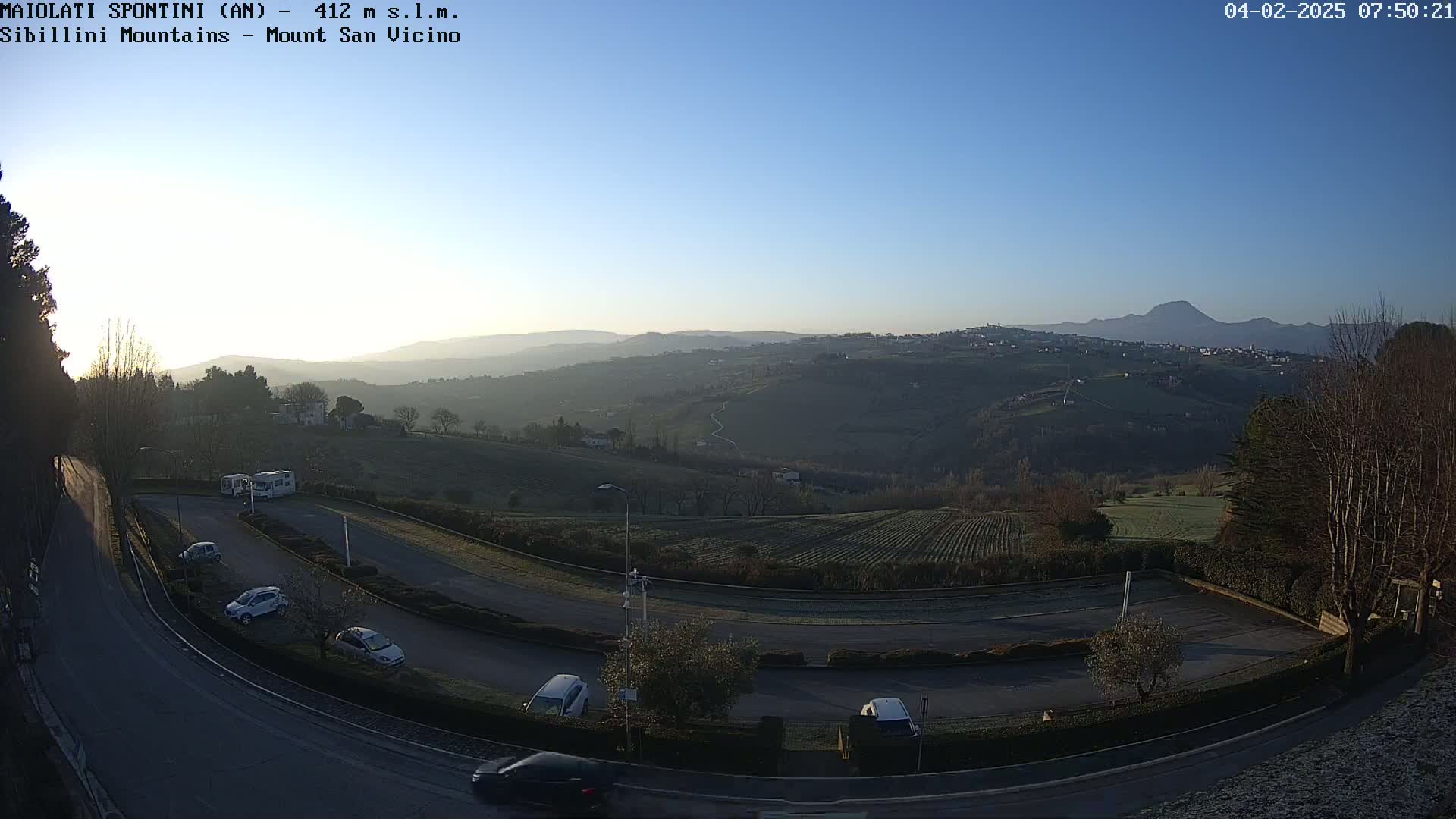 A clear, sunrise view shows a winding road in a parking area overlooking a valley with hills and a distant mountain range.
