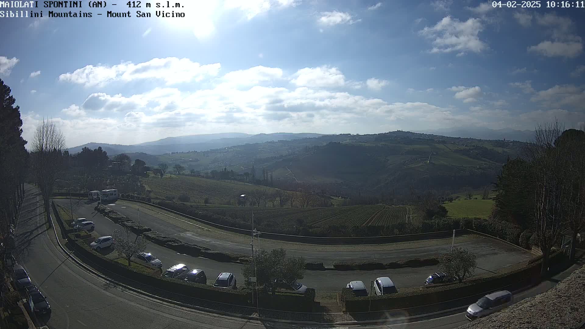 A hilly landscape with vineyards and a parking lot, viewed on a partly sunny day with some clouds.