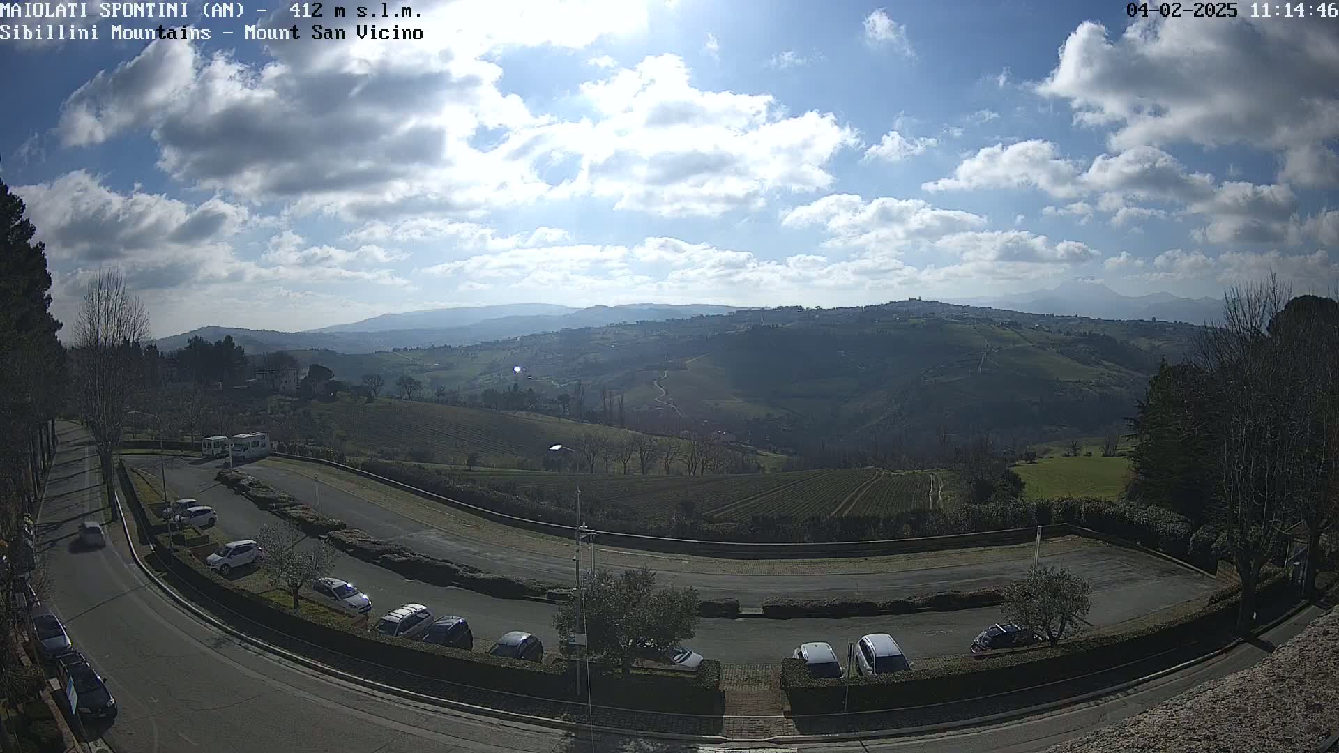A partly cloudy day reveals a winding road with parked cars leading to a scenic overlook of rolling hills and distant mountains.