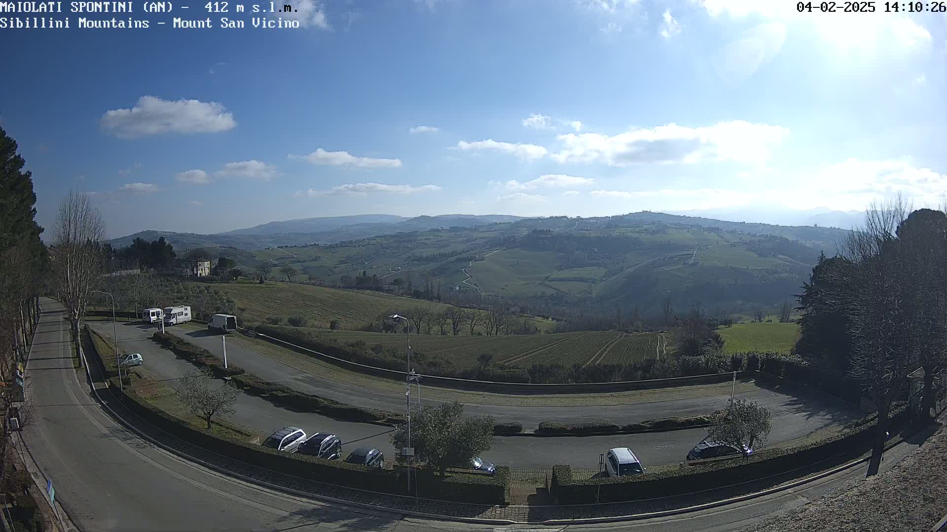 A sunny day reveals a sweeping vista of rolling hills and farmland, overlooking a curved roadway with parked cars and recreational vehicles.