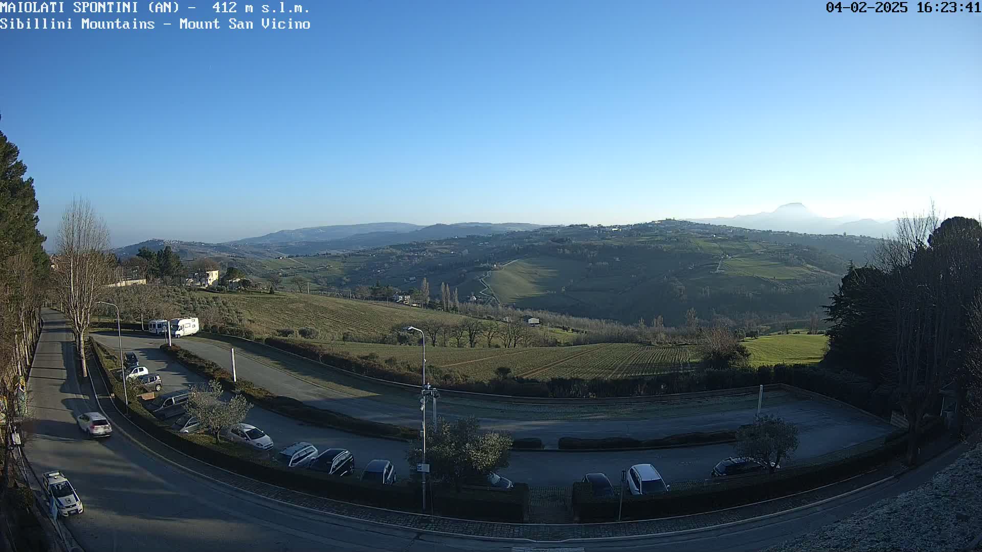 A sunny, clear day reveals a parking lot overlooking rolling hills and distant mountains, with several cars parked and a few trees scattered throughout the landscape.