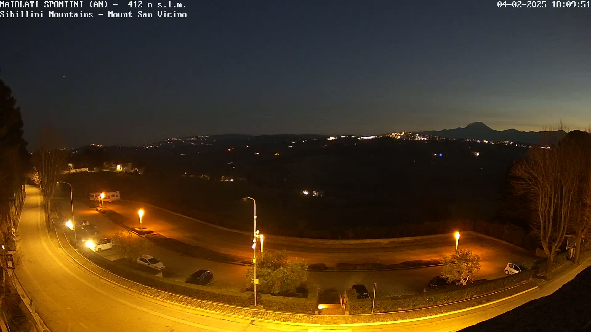 A nighttime, overhead view of a curving road with parked cars and illuminated streetlights, overlooking a distant town nestled in dark hills under a clear, twilight sky.