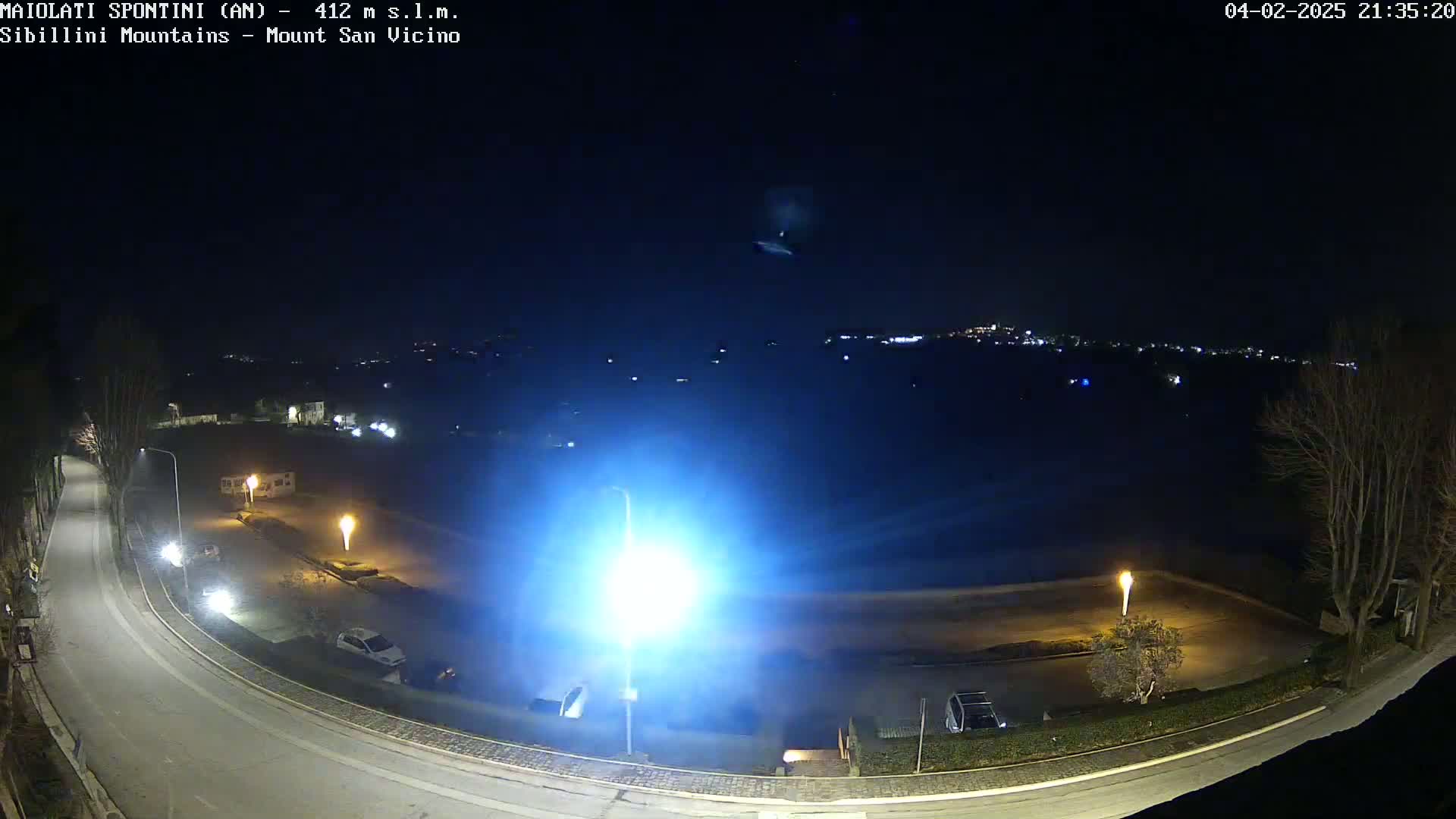 A nighttime, high-angle view of a curving road with parked cars and lit streetlamps, overlooking a dark, empty parking lot and a distant city skyline under a clear sky.
