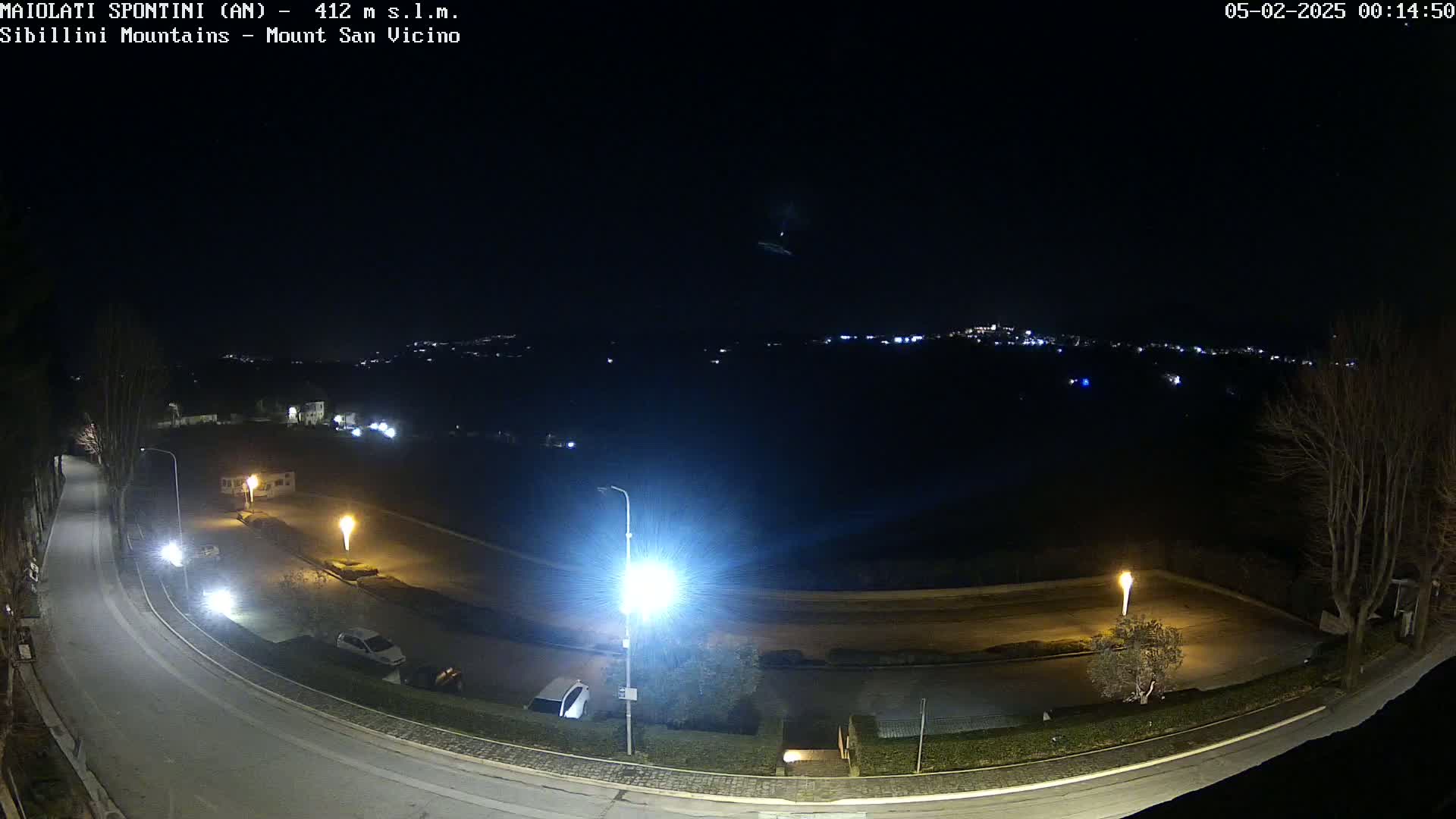 A nighttime, high-angle view shows a curving road illuminated by streetlights, a parking lot with a few cars, and a distant town glowing faintly under a clear, dark sky.