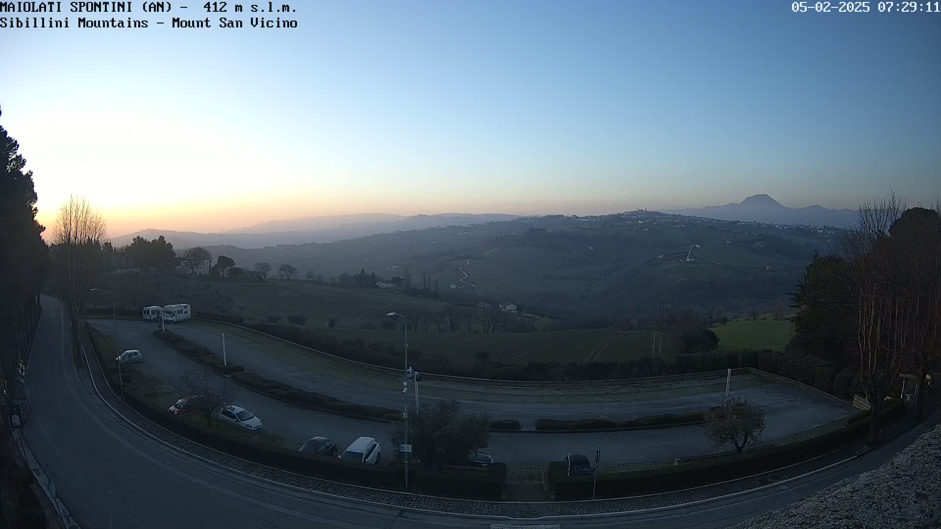 A high-angle, long shot shows a parking lot with several cars and RVs situated on a curved road overlooking a hilly landscape at dawn under a clear, twilight sky.