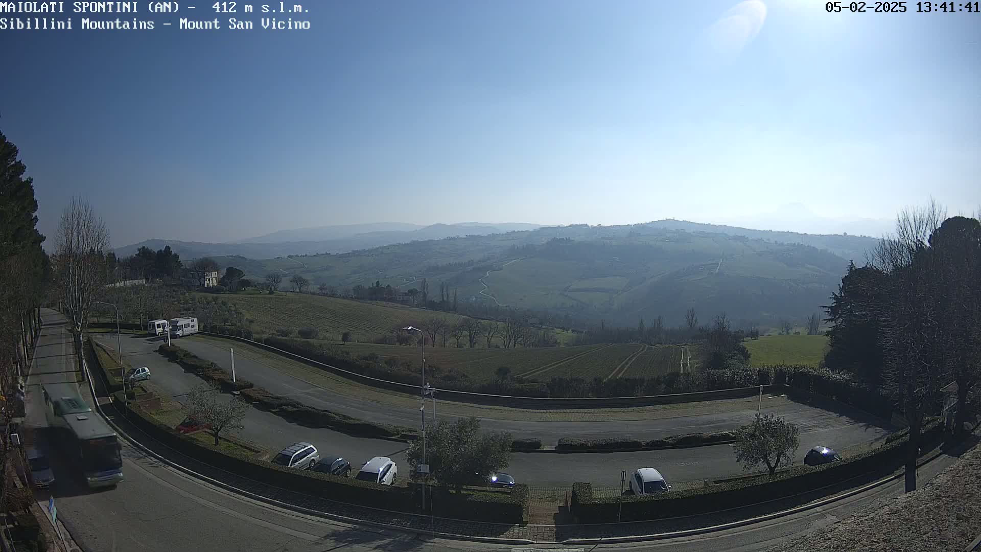 A sunny, clear day overlooks a winding road with parked cars and a bus, leading to a vast expanse of rolling green hills and mountains in the distance.