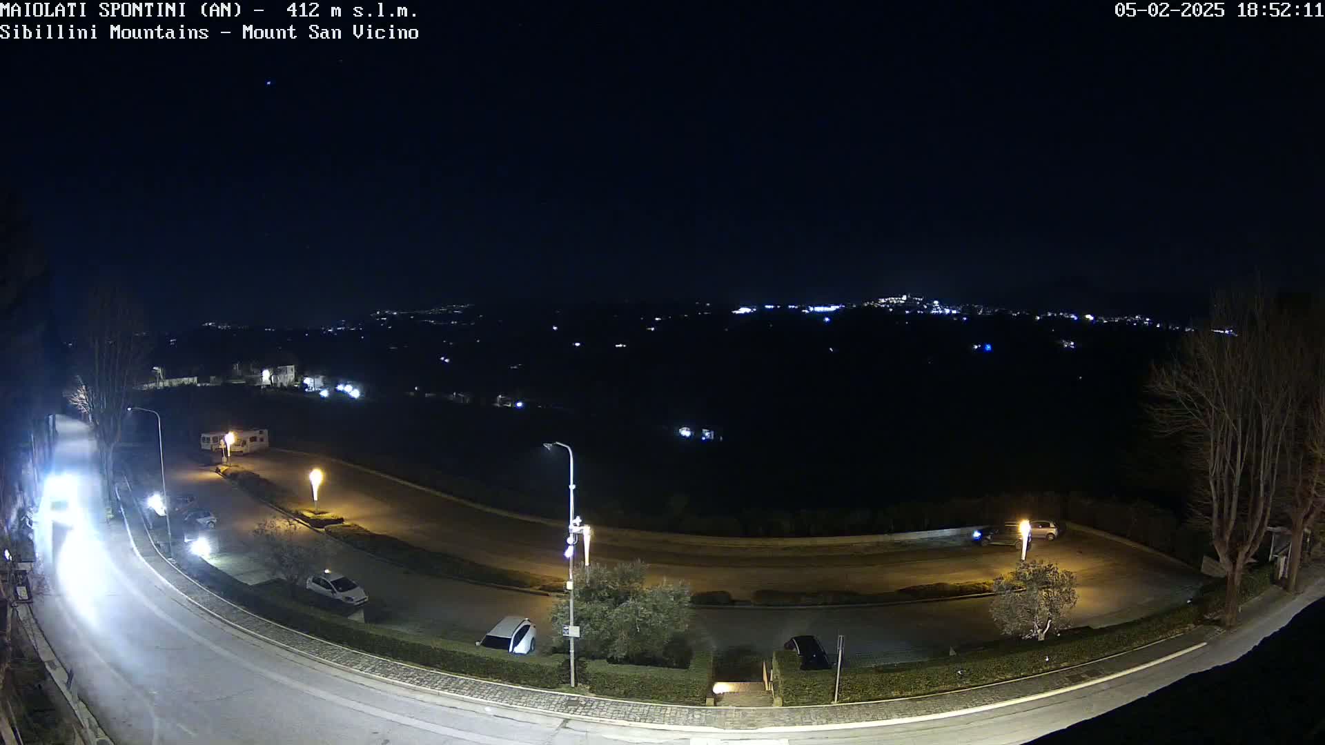 A nighttime, high-angle view of a curving road with parked cars and a distant town illuminated under a clear, dark sky.