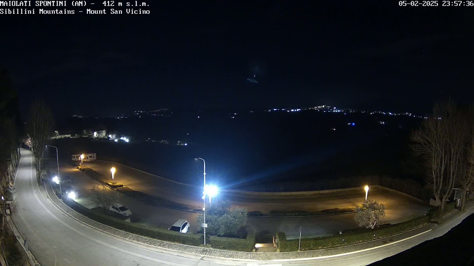 A nighttime, overhead view of a curving road with parked cars and illuminated by streetlights, overlooking a distant town under a clear, dark sky.