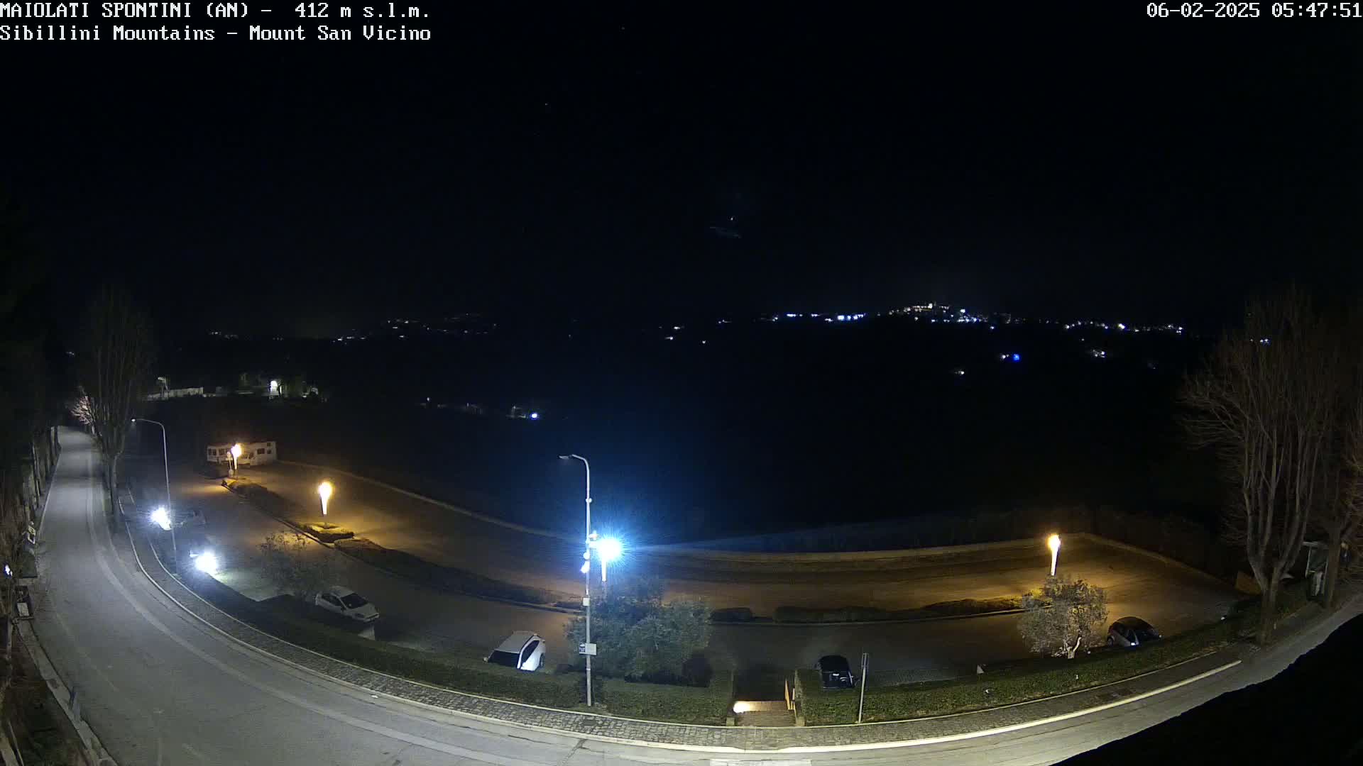 A nighttime, overhead view of a curved road with parked cars and illuminated lamp posts, overlooking a dark, hilly landscape with distant lights.
