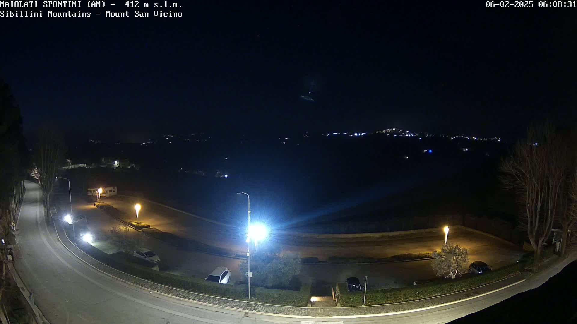 A nighttime, outdoor view of a curving road with parked cars and illuminated lampposts, overlooking a distant town with lights under a clear, dark sky.