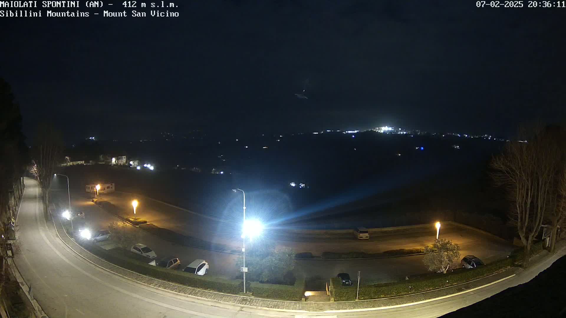 A nighttime, high-angle view of a paved road curving through a parking lot with several cars and light posts, overlooking a distant city illuminated under a dark sky.