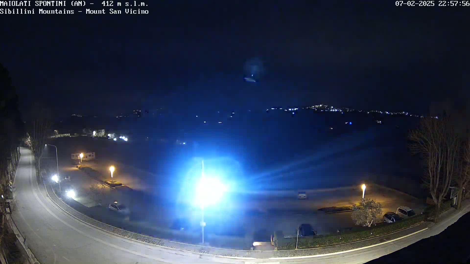 A nighttime view of a mostly empty parking lot illuminated by bright lights, a curved road, and sparsely-leafed trees, with a distant town or city glowing faintly in the background under a dark, clear sky.