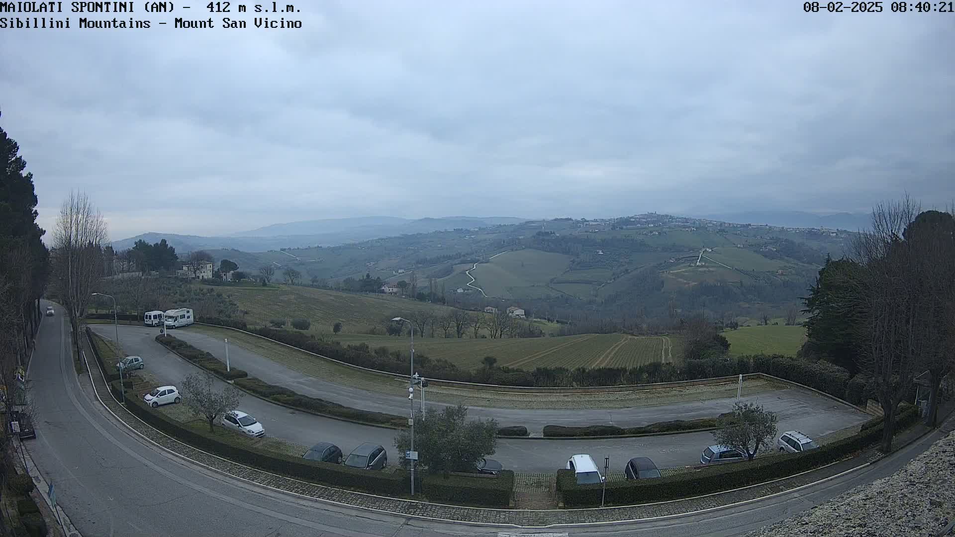 A paved parking lot curves around a hilltop overlooking a valley of rolling green hills under an overcast sky.