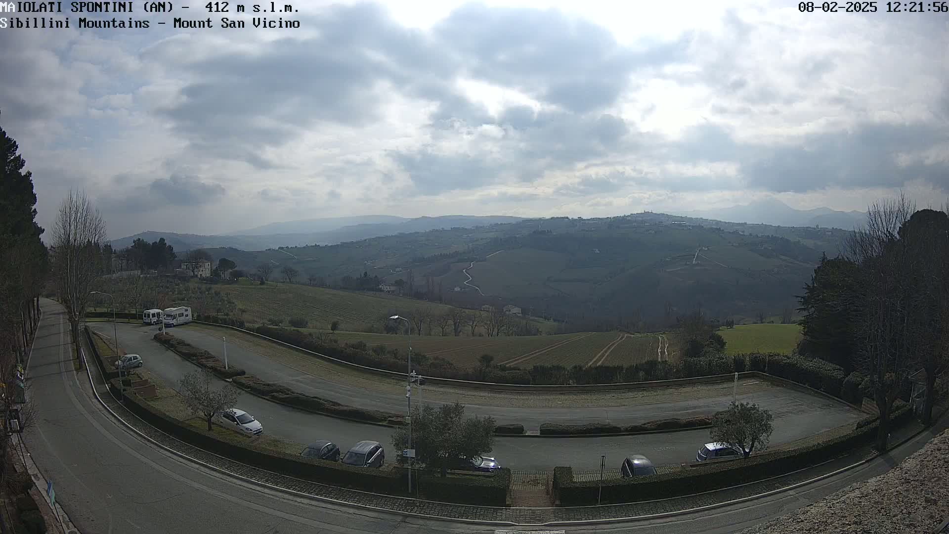 A mostly cloudy day overlooks a paved parking lot with several cars and RVs, situated on a curved road with a view of rolling hills and distant mountains.