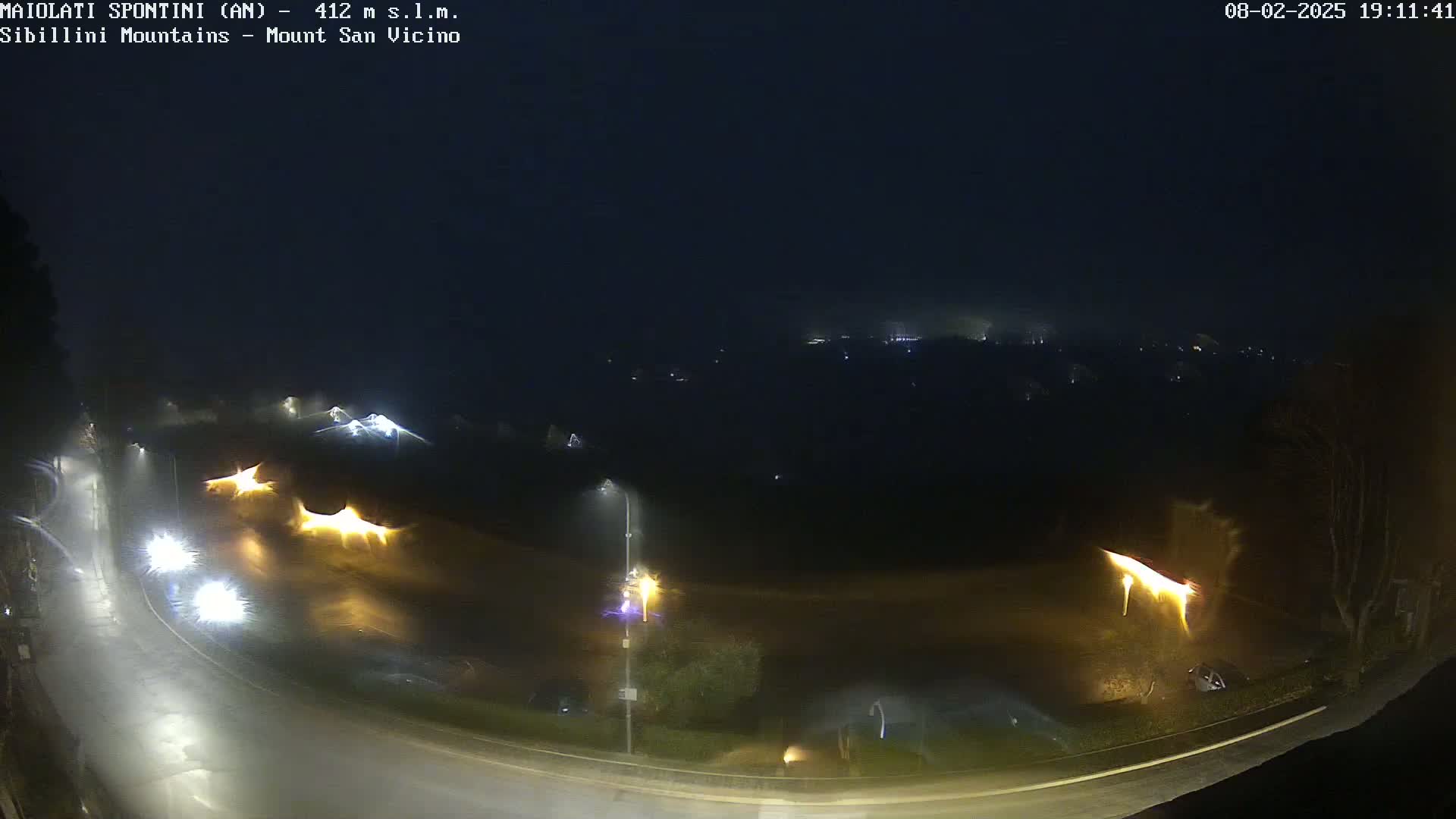 A nighttime, rainy scene shows a curving road with illuminated streetlights and a few vehicles, overlooking a dark, foggy landscape with distant lights.