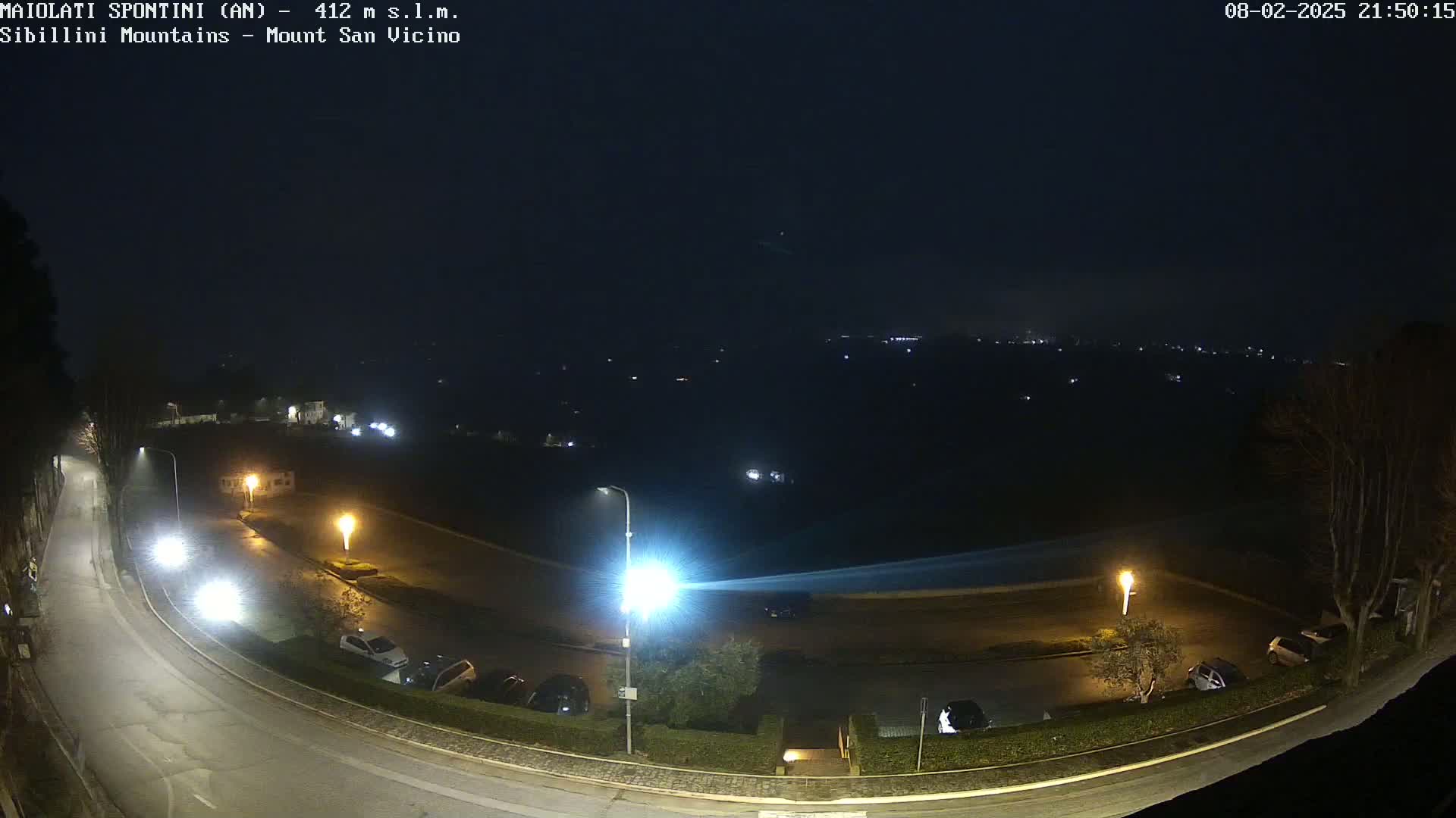 A nighttime, dimly lit parking lot and road curve are seen, overlooking a dark landscape with distant lights under a hazy sky.
