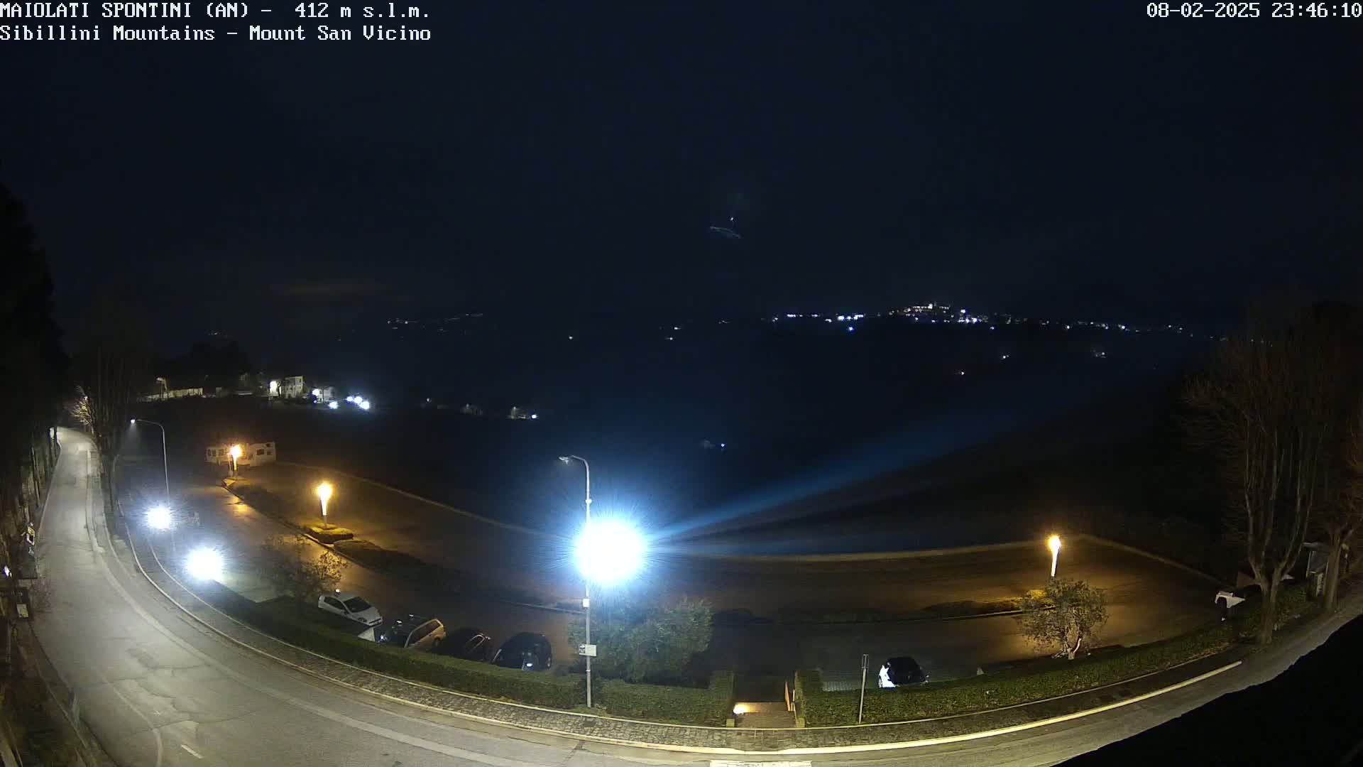 A nighttime, dimly lit parking lot and curving road are visible, with a distant town glowing faintly under a dark, possibly cloudy sky.