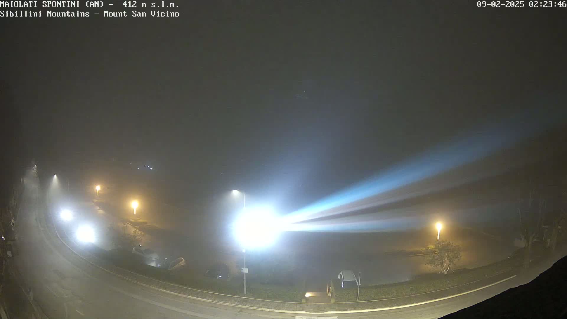 A foggy night scene shows a curving road with parked cars and illuminated streetlights, with a bright light beam cutting across the scene.