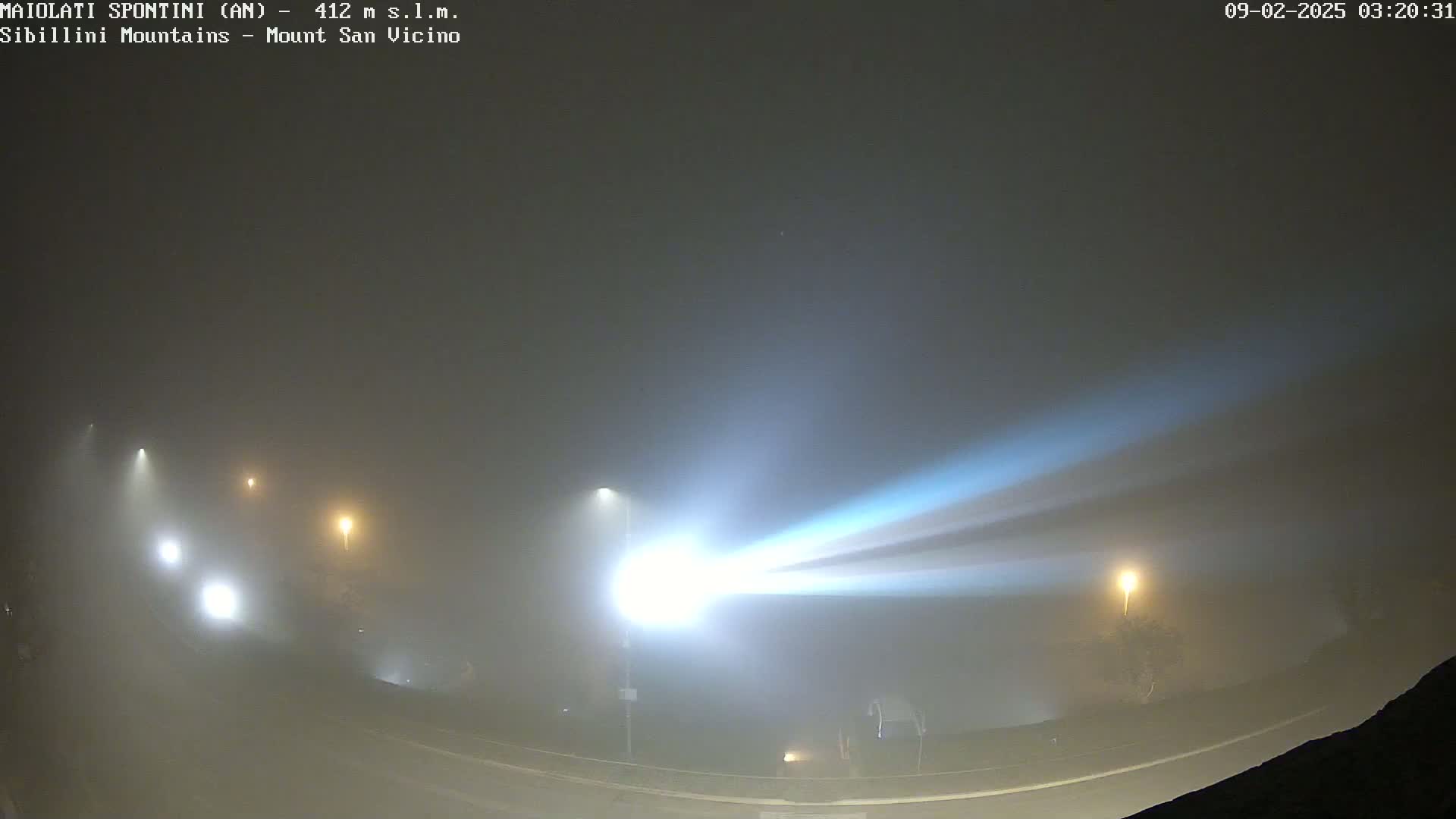 Bright beams of light cut through a dense fog at night, illuminating a road with several streetlights.