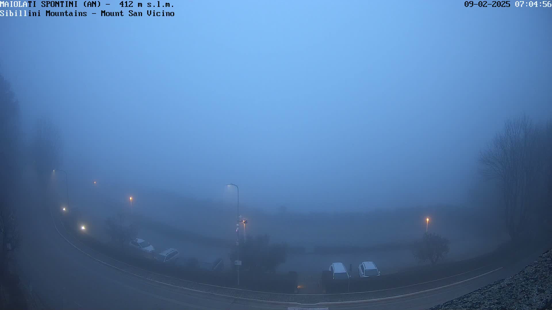 A foggy, dimly lit road curves through a parking lot with several cars parked, under a pale blue sky.