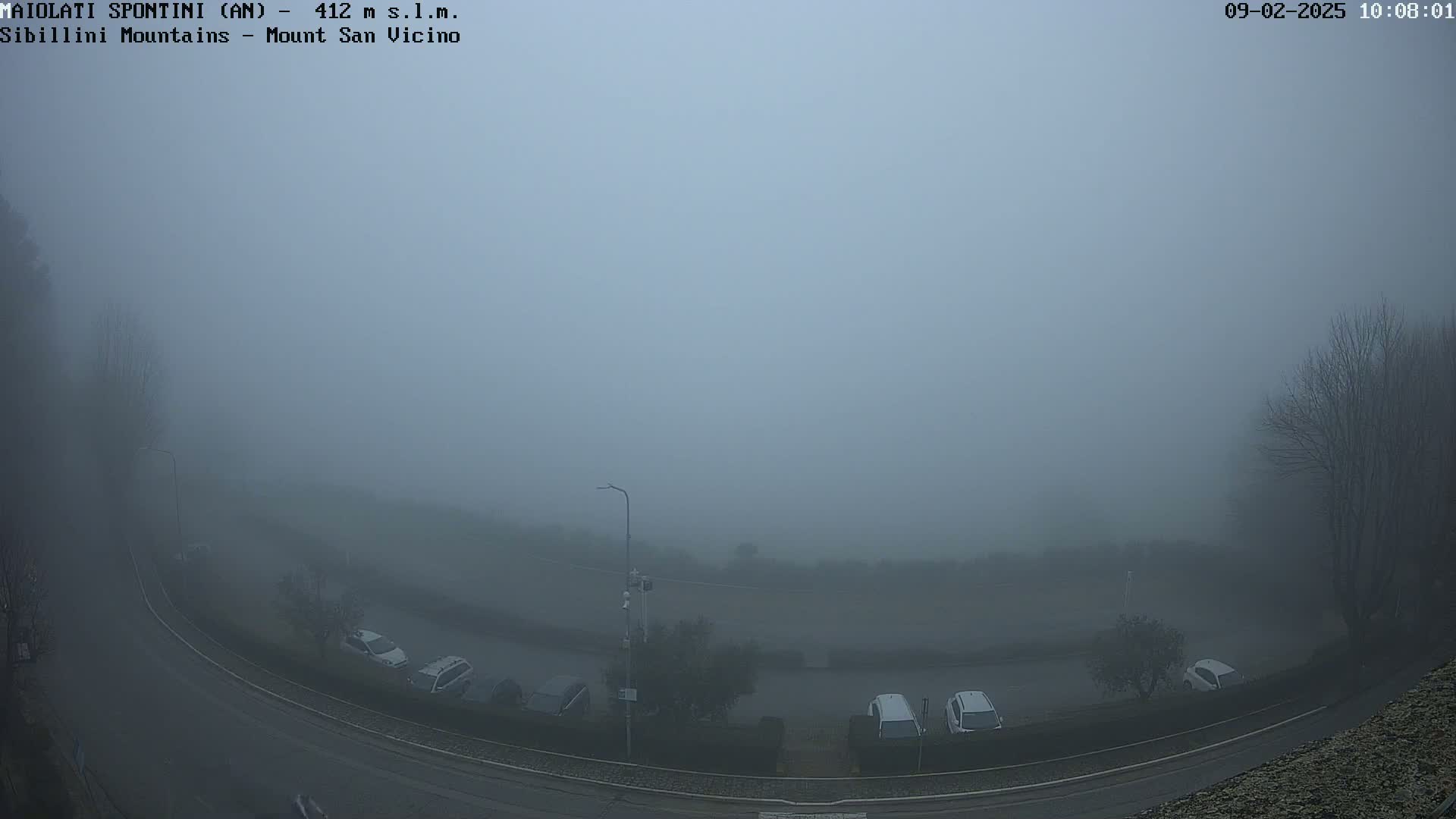 A foggy overlook shows a road curving through a parking lot with several cars parked, all obscured by thick fog.