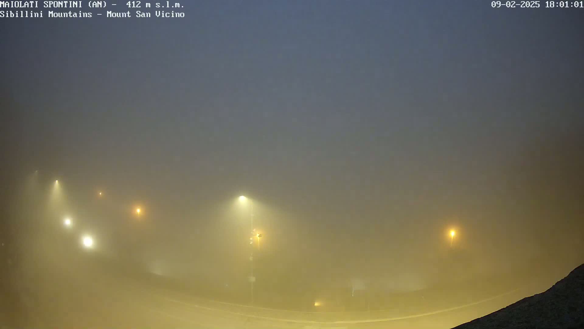 A foggy night scene shows a road with several illuminated streetlights, and some faint vehicle lights visible through the dense fog.