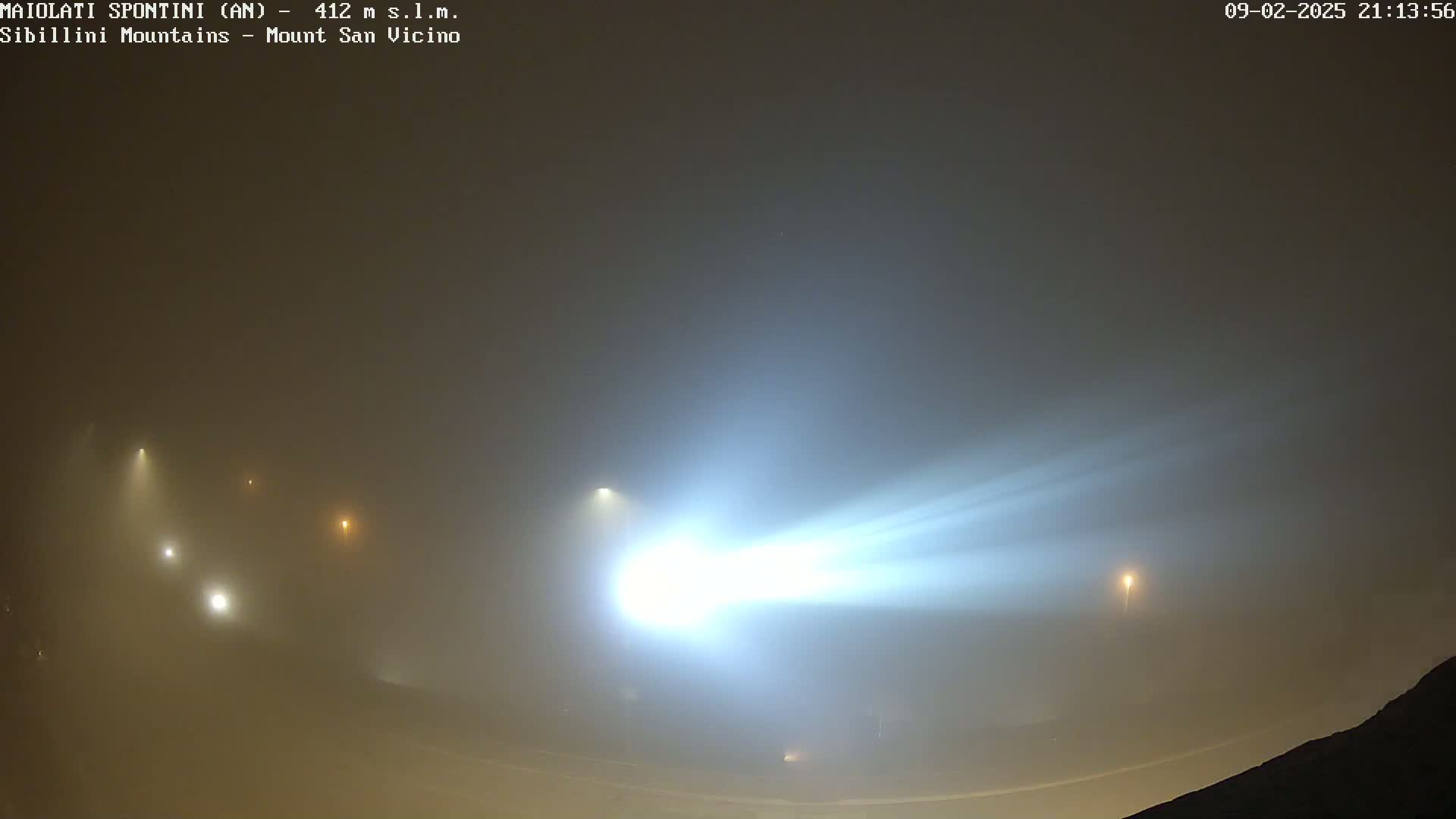 A bright light cuts through a thick fog at night, illuminating several dimly lit streetlights in the distance.