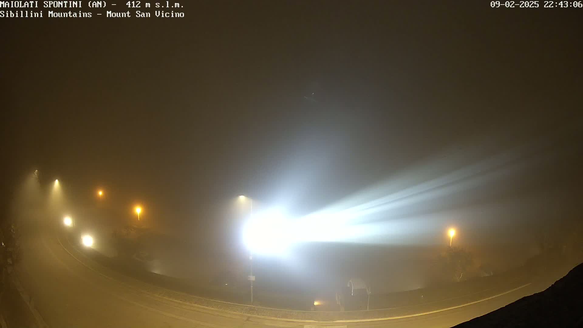 A bright light cuts through heavy fog illuminating a curved road with a few vehicles and streetlights at night.