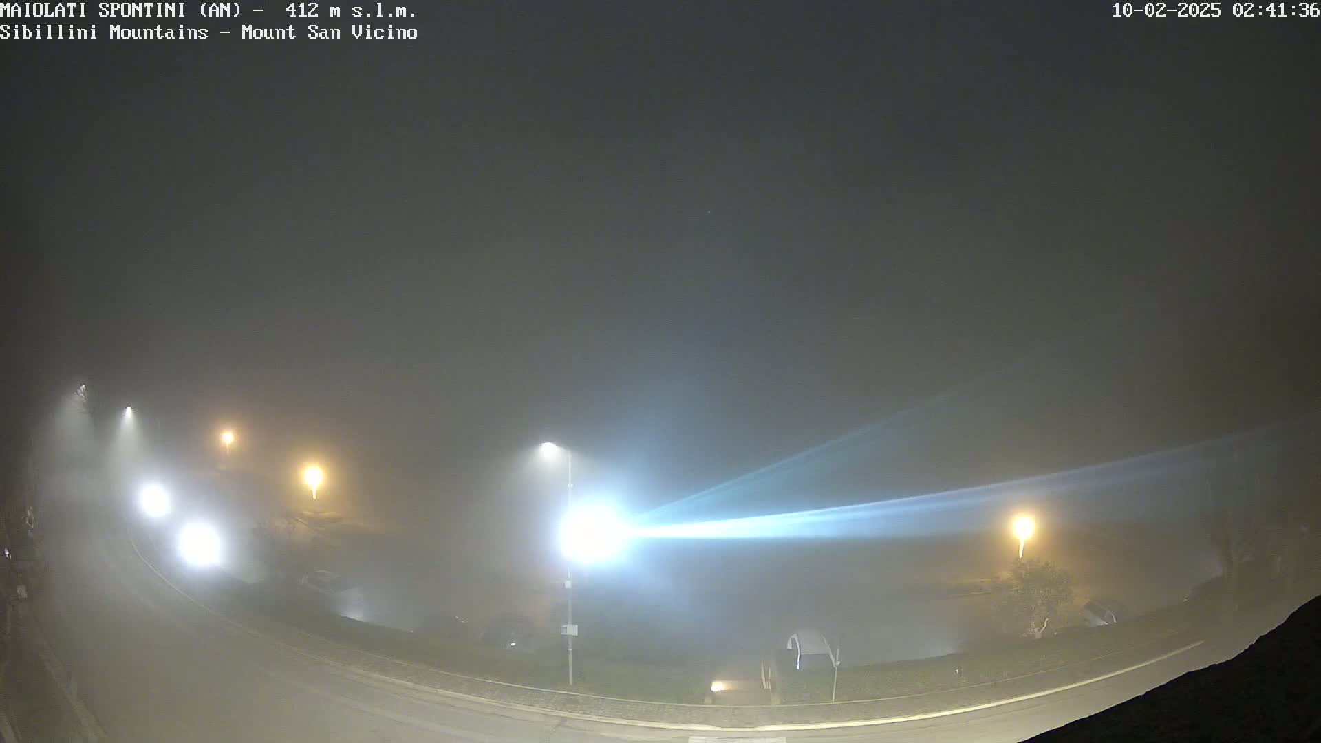 A foggy night scene shows a curved road with several cars and streetlights visible through the dense fog.