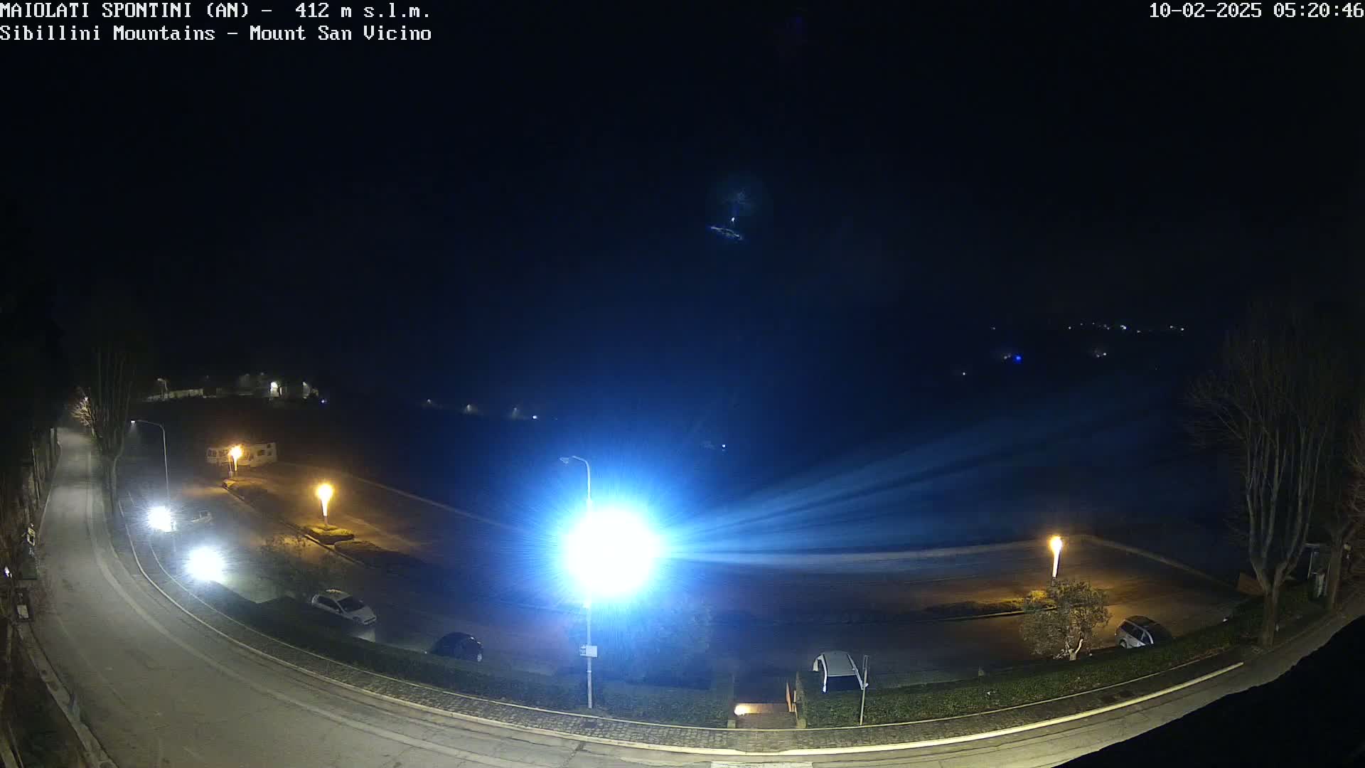 A nighttime, high-angle view of a curving road with parked cars and illuminated streetlights, overlooking a dark, empty parking lot and distant, faintly lit buildings under a clear, dark sky.