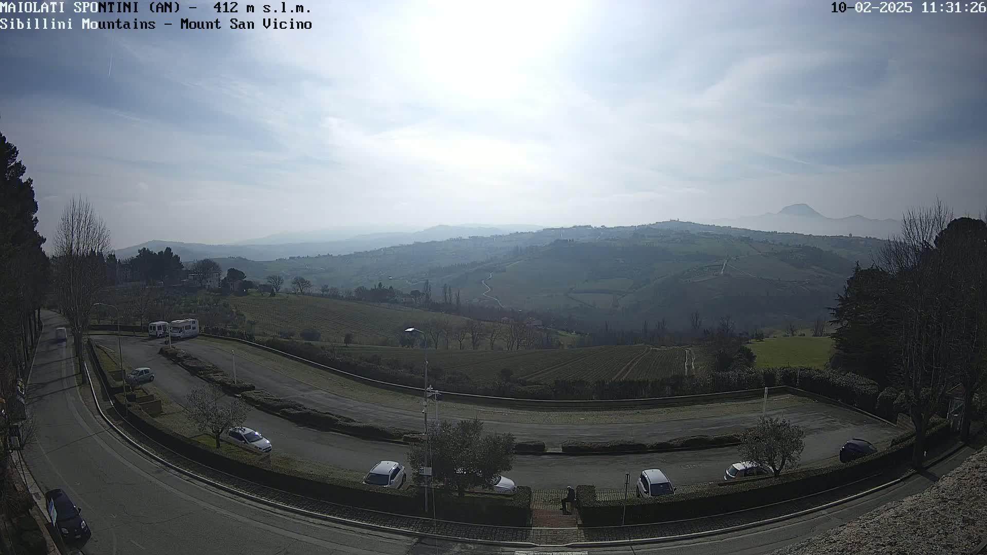 A mostly sunny day reveals a sweeping view of rolling hills, vineyards, and a parking lot with several cars, all overlooking a distant mountain range.
