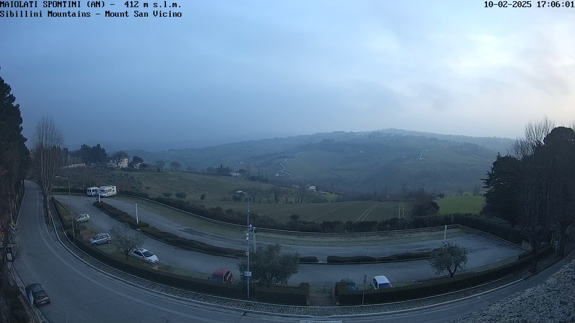 An empty parking lot overlooks a hazy valley of rolling hills and farmland under a pale blue sky.