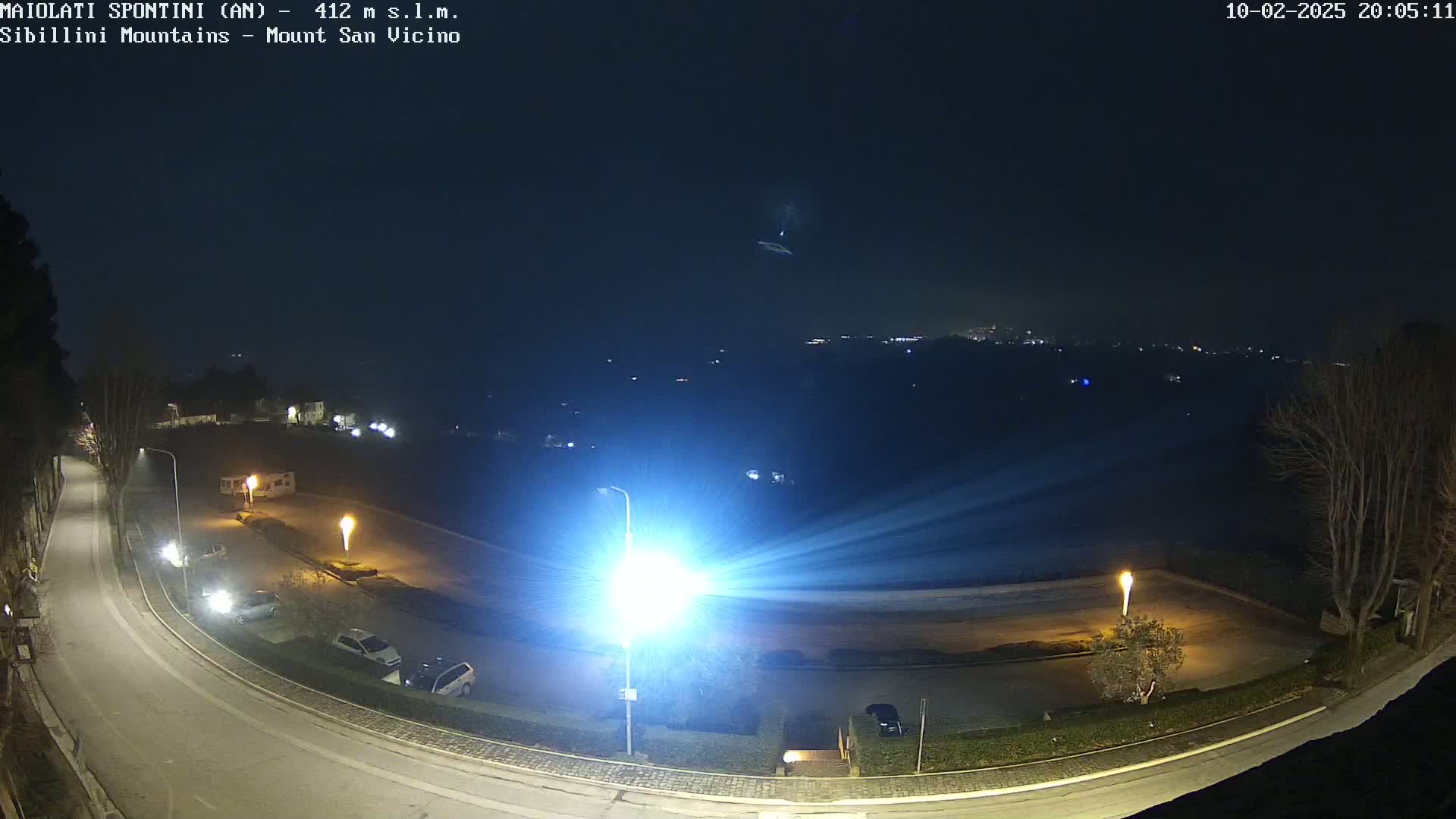 A nighttime, high-angle view of a mostly empty parking lot and curving road illuminated by bright lights, with a distant cityscape visible under a dark, clear sky.