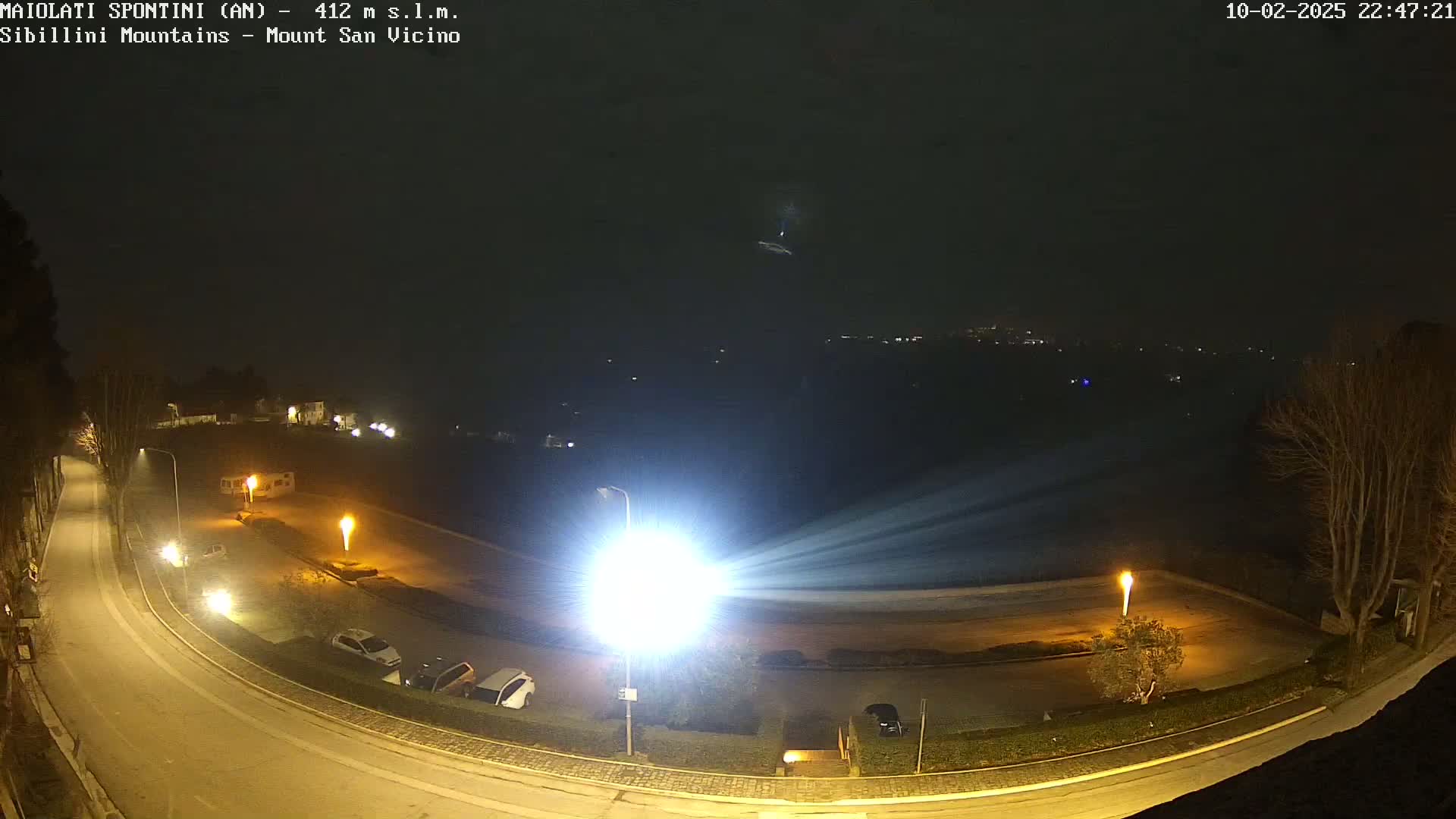 A nighttime view of a parking lot and curved road illuminated by bright lights, with a hazy sky and bare trees in the background.